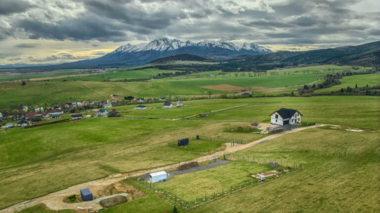Panoramatický pohľad na vidiecku krajinu s domom a vzdialenými zasneženými horami pod zamračenou oblohou.